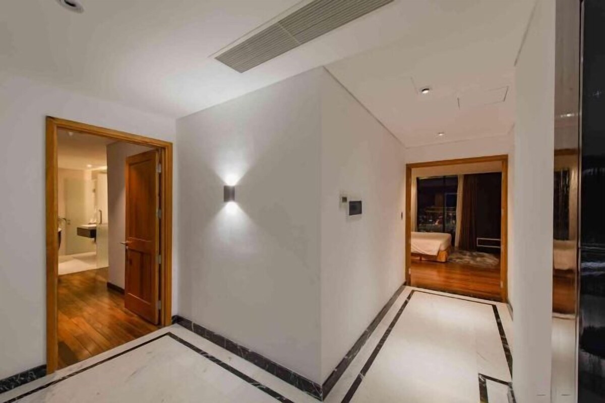 Un couloir d’hôtel avec une porte en bois donnant sur une salle de bain, un luminaire fixé au mur et une chambre avec un lit et des rideaux.
