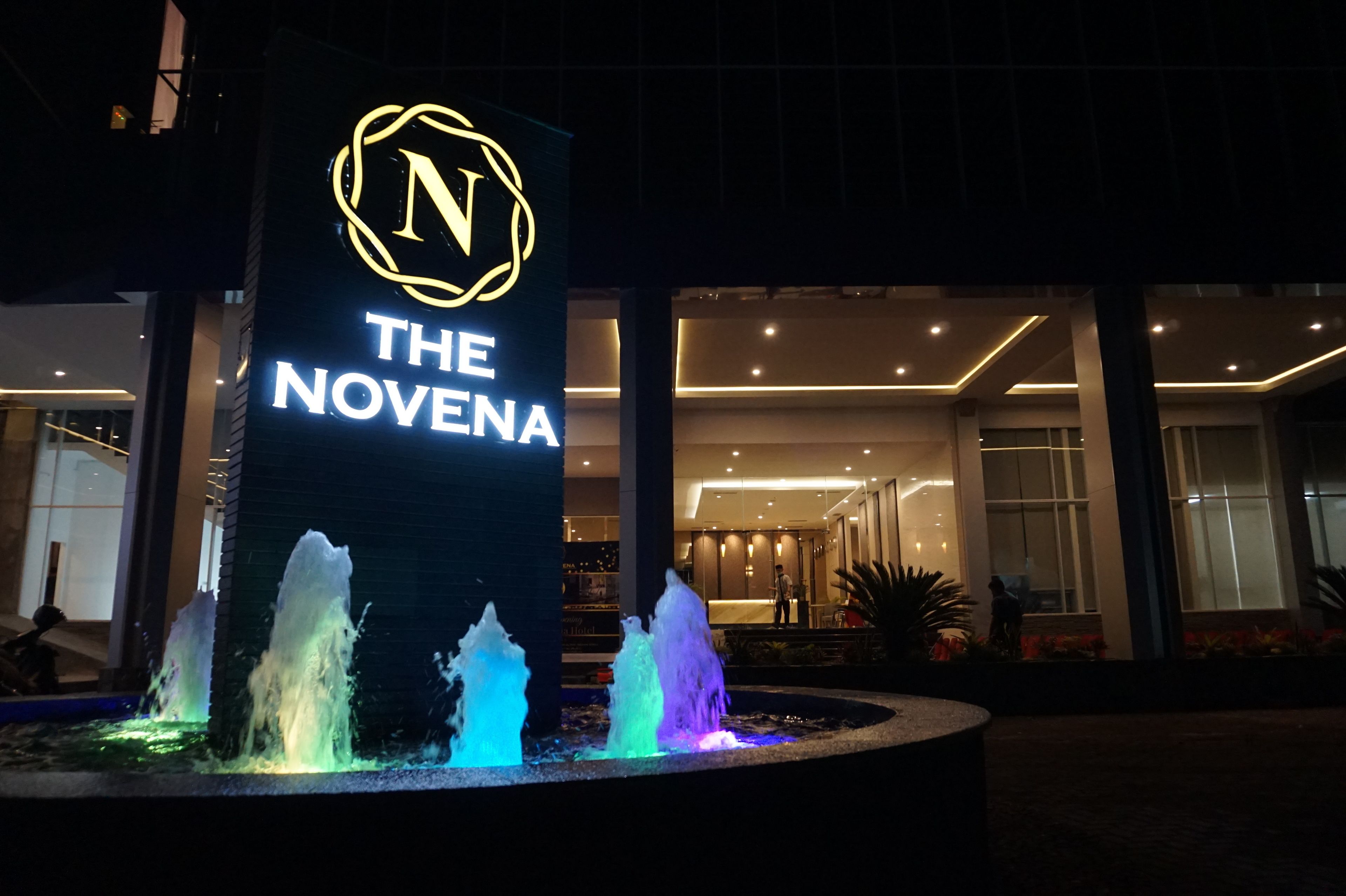 A hotel entrance with a neon sign reading "THE NOVENA" and a water feature with multiple jets.