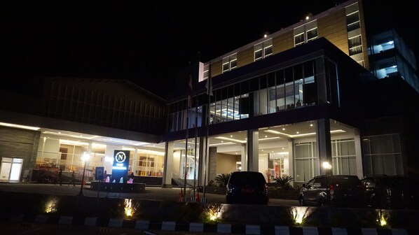 A modern building with a glass facade, a visible entrance, and a parking area with cars.