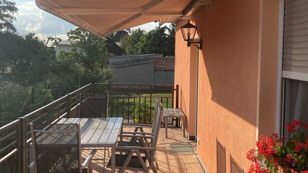 Een terras buiten met een tafel, stoelen, een grote parasol en een bloempot.