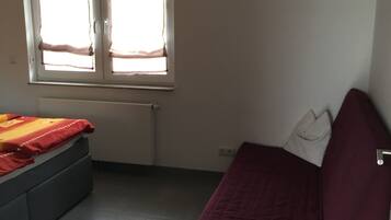 Een kleine, eenvoudig ingerichte kamer met een bed, een slaapbank en een raam met gordijnen.