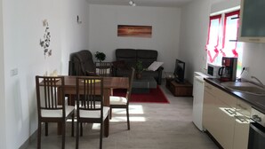 Een moderne woonkamer met een bank, fauteuils en een eettafel. Er is een keukenhoek met een magnetron, koffiezetapparaat en kastjes.