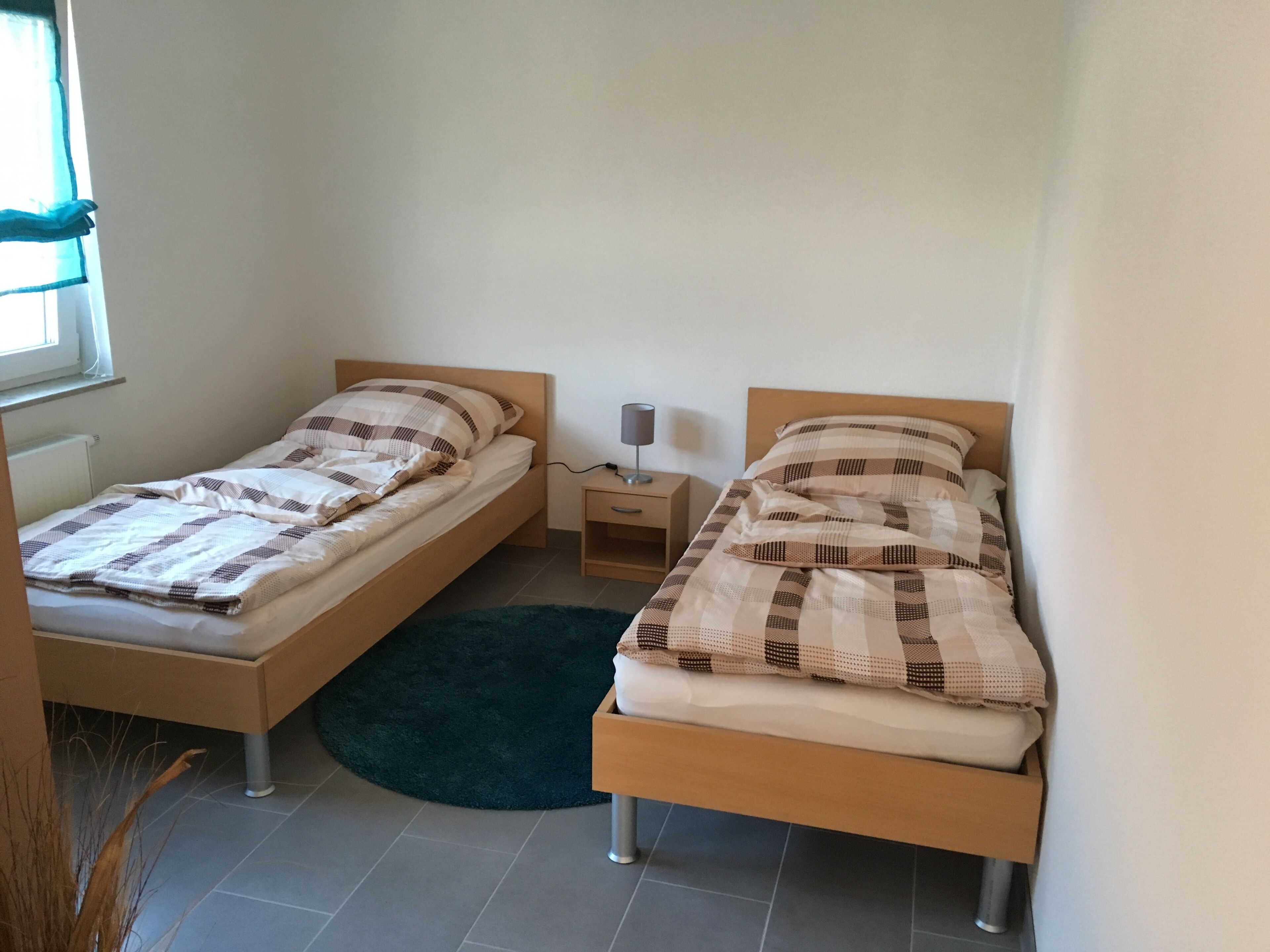 Ein kleines Zimmer mit zwei Einzelbetten, einem Nachttisch mit Lampe, einem dunklen Teppich und einem Fenster mit durchsichtigen Vorhängen.