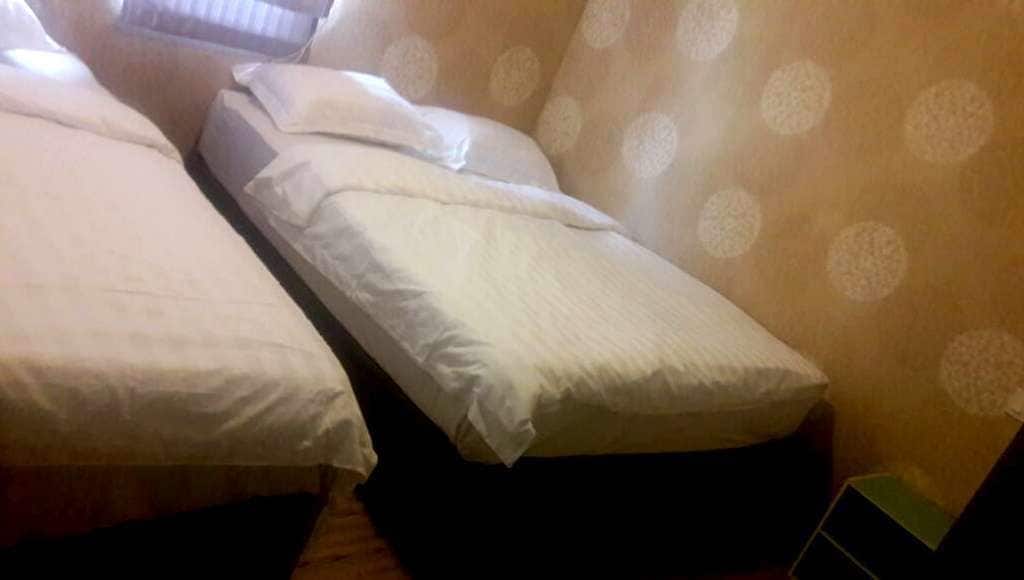 A room with two single beds, each with white bedding and pillows. The room has a patterned wall and a wooden floor.