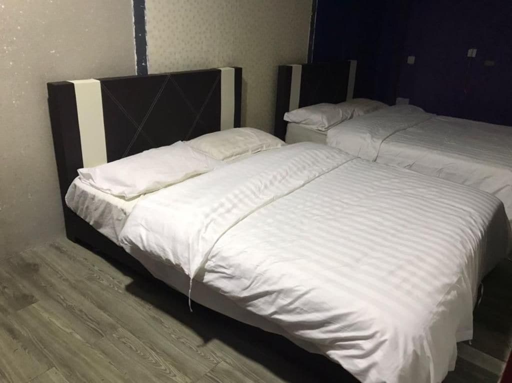 A room with two beds, each with white bedding and headboards. The room has a wooden floor and a dark-colored wall.