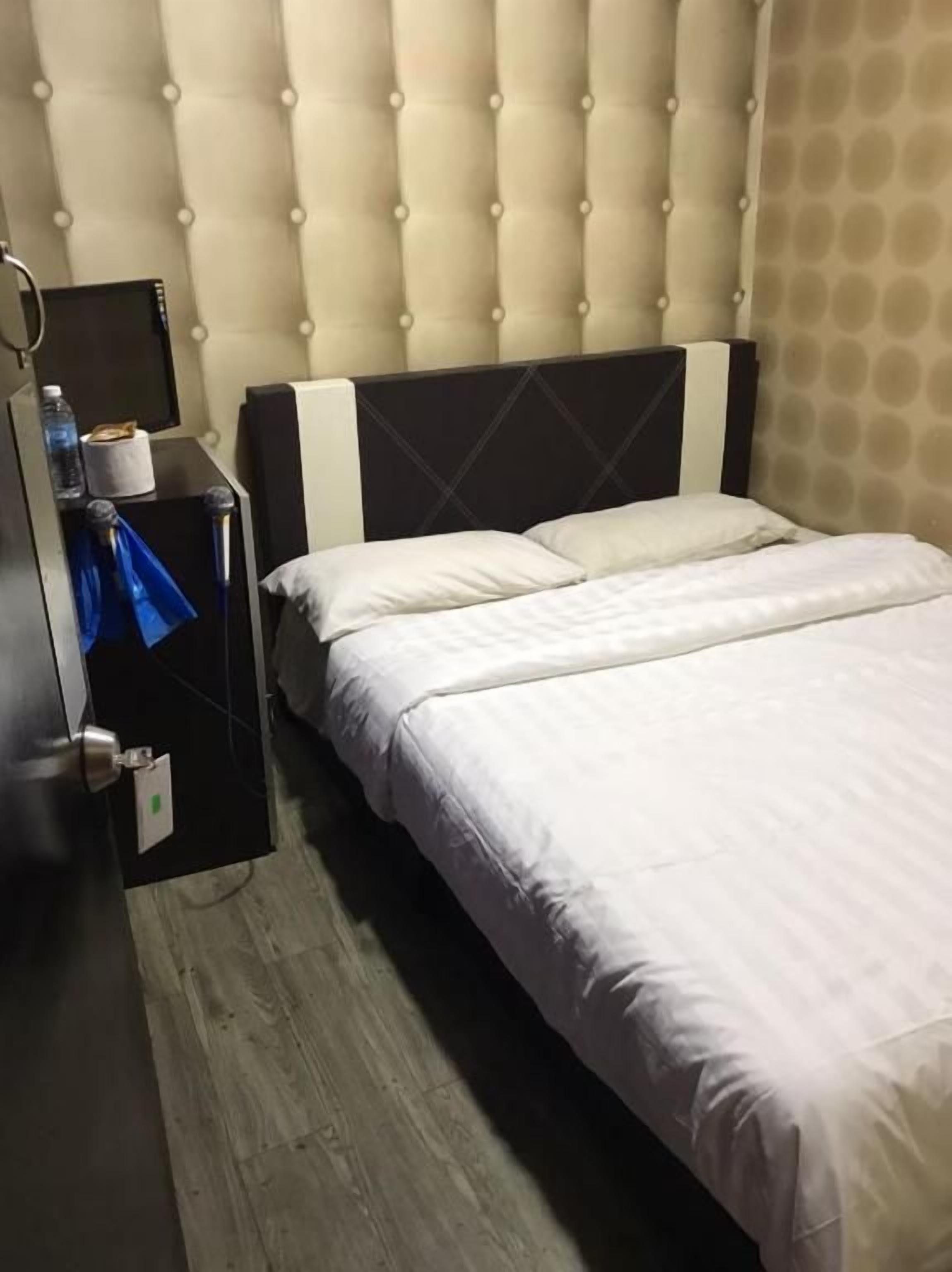 A hotel room with a tufted headboard, a bed with white linens, a nightstand with a lamp, and a wall-mounted TV.
