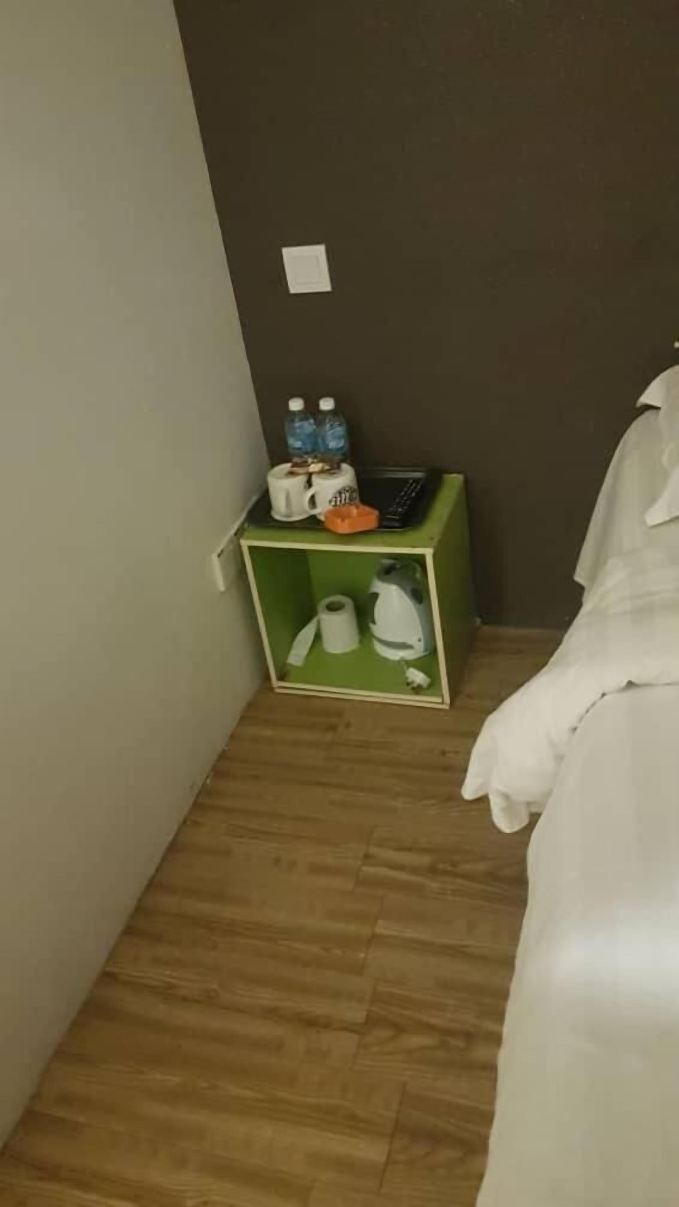 A corner of a room with a green storage box containing a kettle, a roll of toilet paper, and two bottles of water.