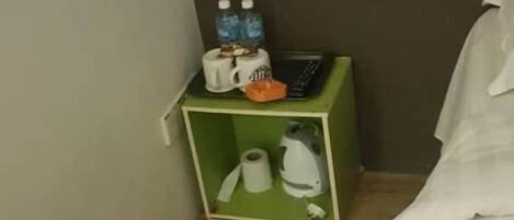A corner of a room with a green storage box containing a kettle, a roll of toilet paper, and two bottles of water.