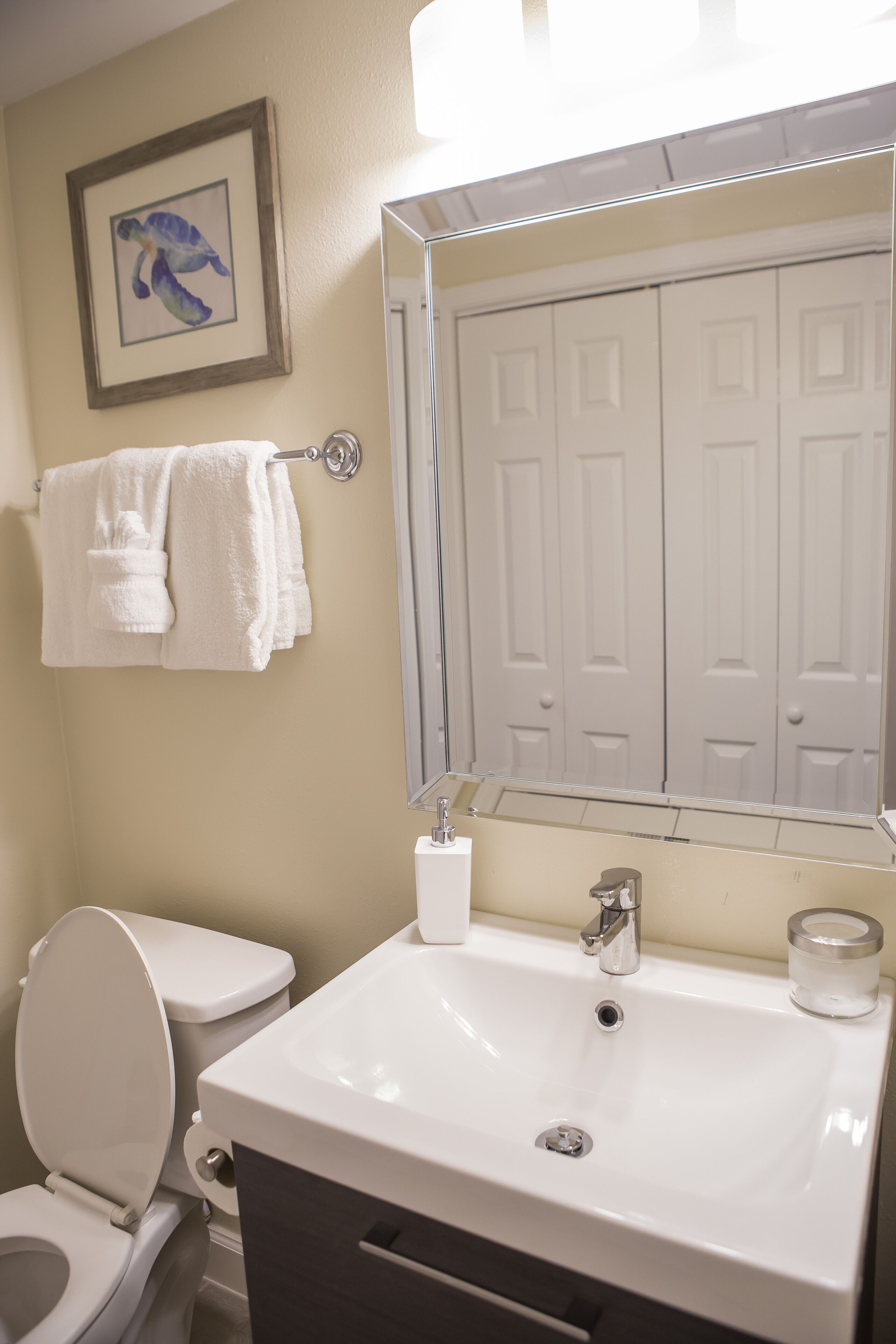 A bathroom with a white sink, mirror, and toilet. There is a framed picture on the wall and a towel rack with towels.