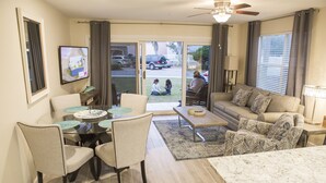 A modern living room with a dining area, a sofa, and a TV mounted on the wall. - NewBestChoice🌴2pools,1Hot, Internet,Tennis🌞BeachSvc,2 Chair,1 Umbrella,Sleep 6 (Miramar Beach)