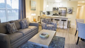 A modern living room with a sofa, armchairs, a coffee table, and a kitchen area with a refrigerator, microwave, and bar stools. - NewBestChoice🌴2pools,1Hot, Internet,Tennis🌞BeachSvc,2 Chair,1 Umbrella,Sleep 6 (Miramar Beach)