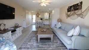 A spacious living room with a ceiling fan, a TV mounted on the wall, a light-colored sofa, and a coffee table. - Jamaica Royale Siesta Key Condo 109 (Siesta Key)