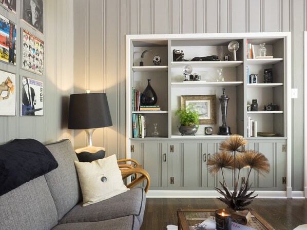 Un salón con un sofá gris, una lámpara negra, una estantería llena de libros y objetos decorativos, y una pared decorada con cuadros.