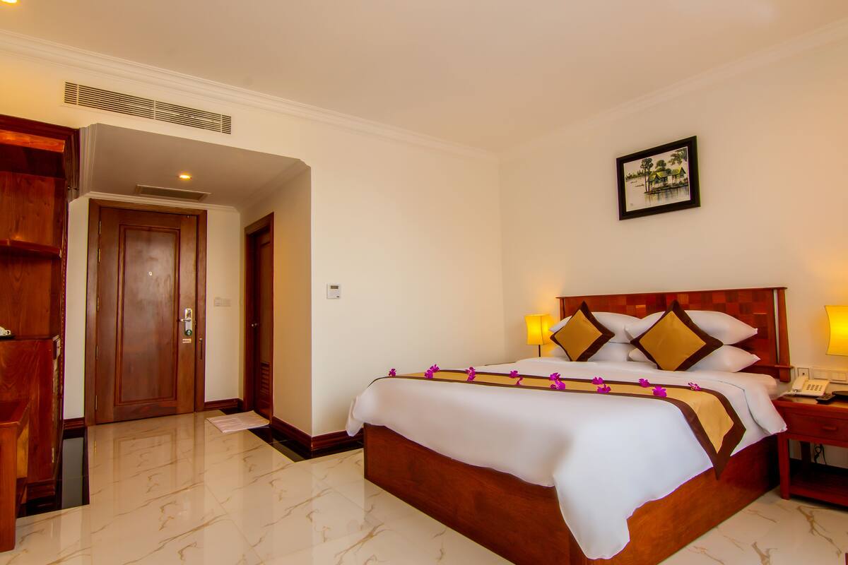 A hotel room with a large bed, wooden headboard, a bedside table with a lamp, and a picture on the wall.