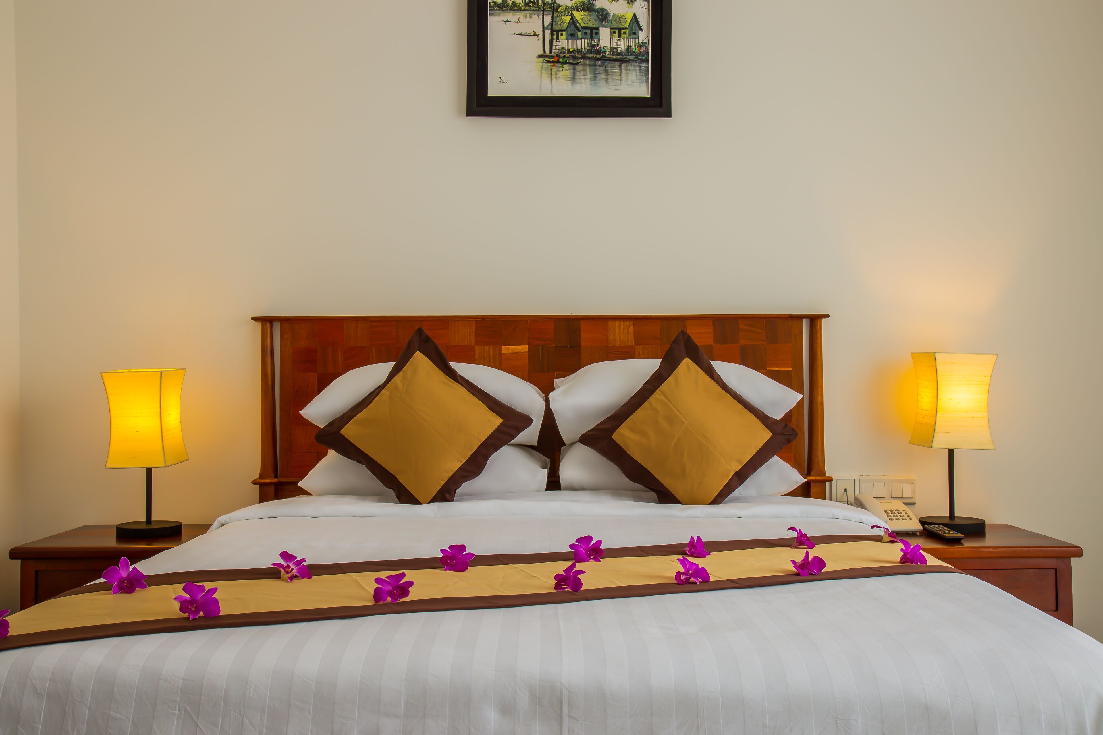 A neatly made bed with white linens, two decorative pillows, and a wooden headboard. There are two bedside tables with lamps and a framed picture on the wall above the bed.