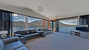 A modern living room with a large sectional sofa, a flat-screen TV, and a view of mountains through large windows. - Amazing Unique Five-Star Waterfront Retreat (Old Beach)