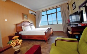 Ein Hotelzimmer mit einem Bett, einem Schreibtisch mit Fernseher, einem Sessel und Blick aufs Meer.