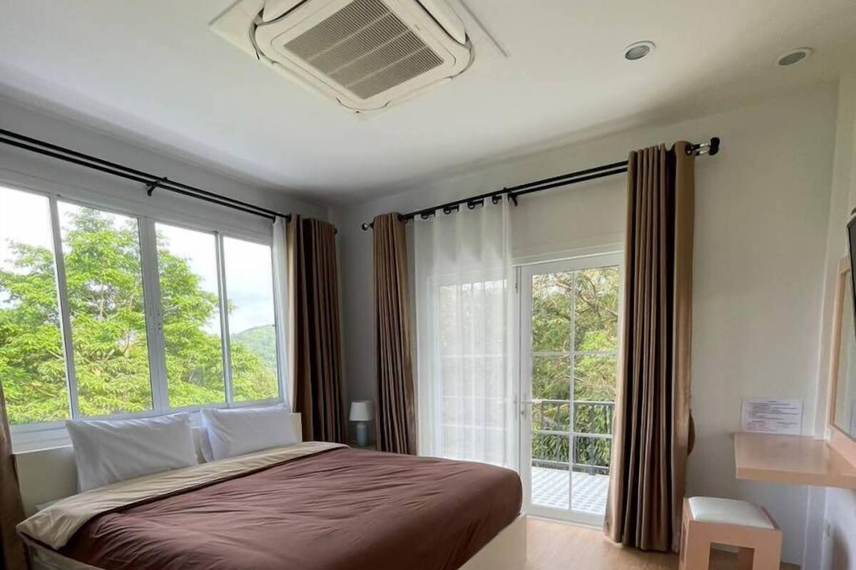 Ein Schlafzimmer mit Bett, einem kleinen Hocker, einem Fenster mit Vorhängen und einem Deckenventilator.