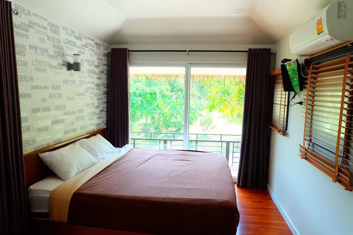 Ein Hotelzimmer mit einem großen Bett, Blick auf Grünflächen, einer Wand mit Städtenamen und einer Klimaanlage.