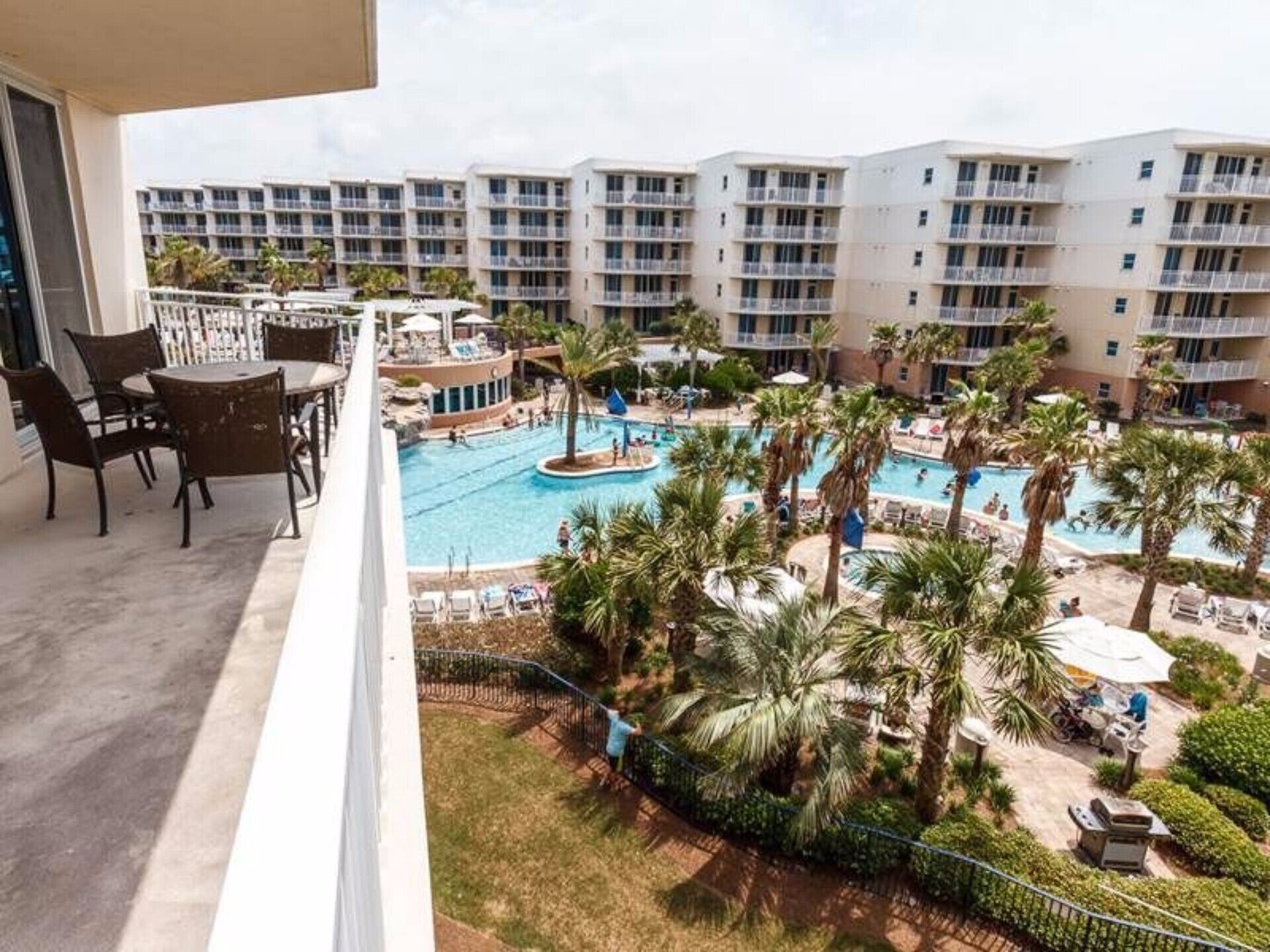 Un balcon d’hôtel donnant sur un espace piscine, comprenant des chaises longues et des palmiers.