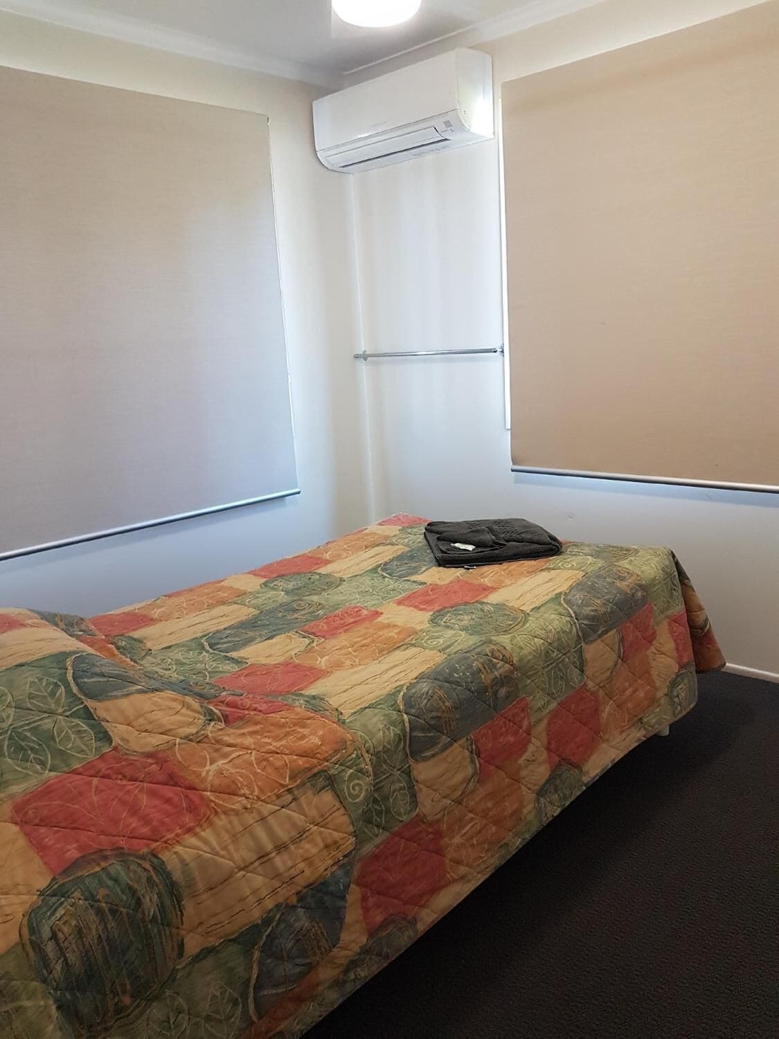 Een kleine eenpersoonskamer met een kleurrijke sprei, een raam met zonwering en een airconditioning op de muur.