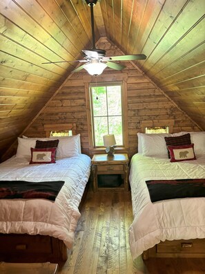 A cozy attic bedroom with two beds, wooden walls, a ceiling fan, and a window with a view of greenery. - Elevate Your Stay! (Crosslake)