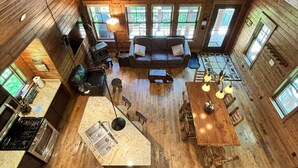 A spacious cabin interior with a high ceiling, wooden walls, multiple windows, a central seating area, a kitchen with a stove, and a dining table. - Elevate Your Stay! (Crosslake)