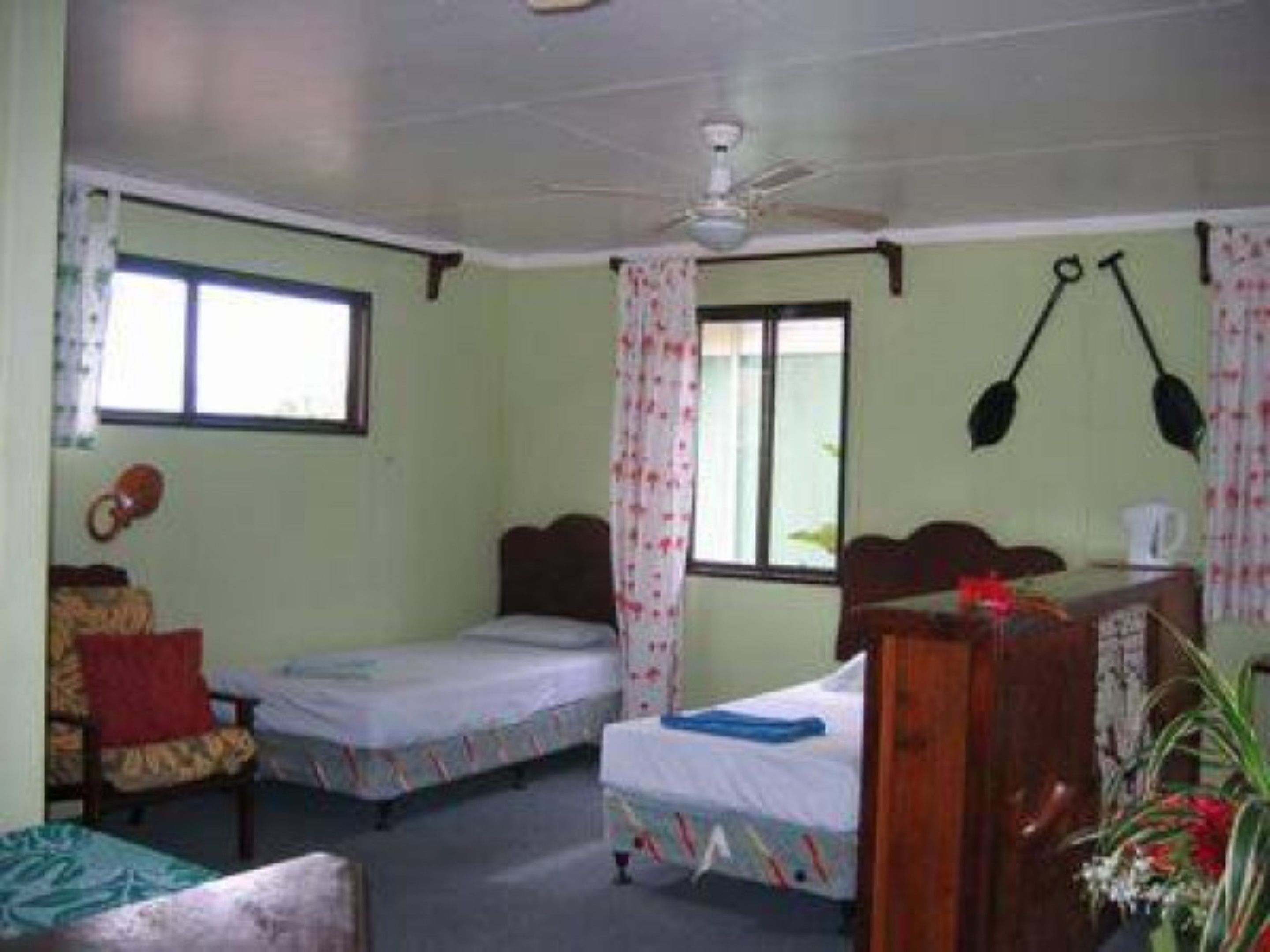 Una habitación pequeña y sencilla con dos camas individuales, un aparador de madera, una silla y un ventilador de techo.