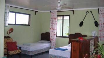 Una habitación pequeña y sencilla con dos camas individuales, un aparador de madera, una silla y un ventilador de techo.