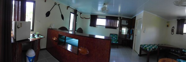 A spacious living area with a wooden reception desk, a large window with curtains, a ceiling fan, and a dining area with a table and chairs.