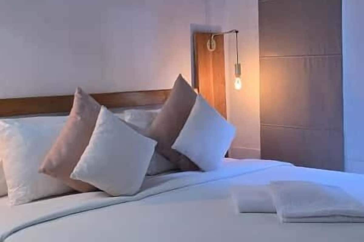 A neatly made bed with white bedding and several pillows, a wooden ceiling, and a wall-mounted air conditioning unit.