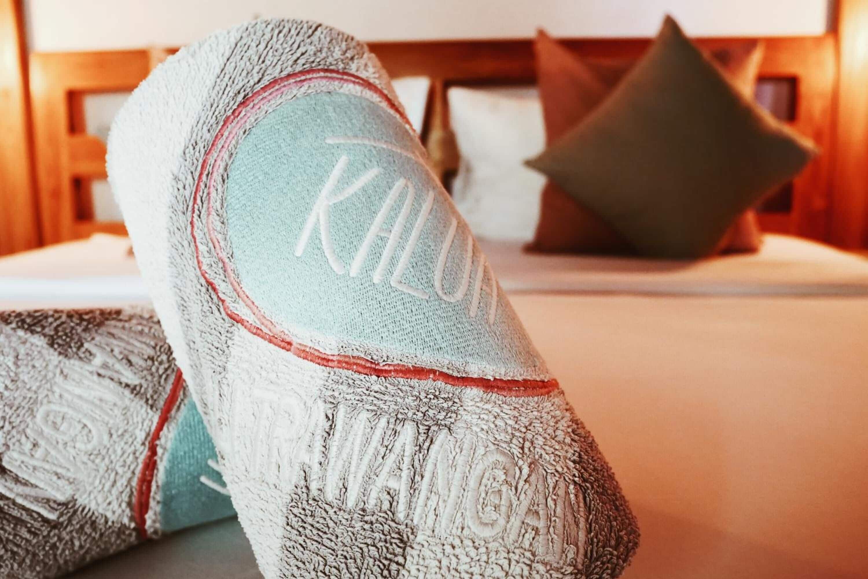 A neatly folded towel with the word "KILUN" embroidered on it, placed on a bed with a wooden headboard and a pillow.