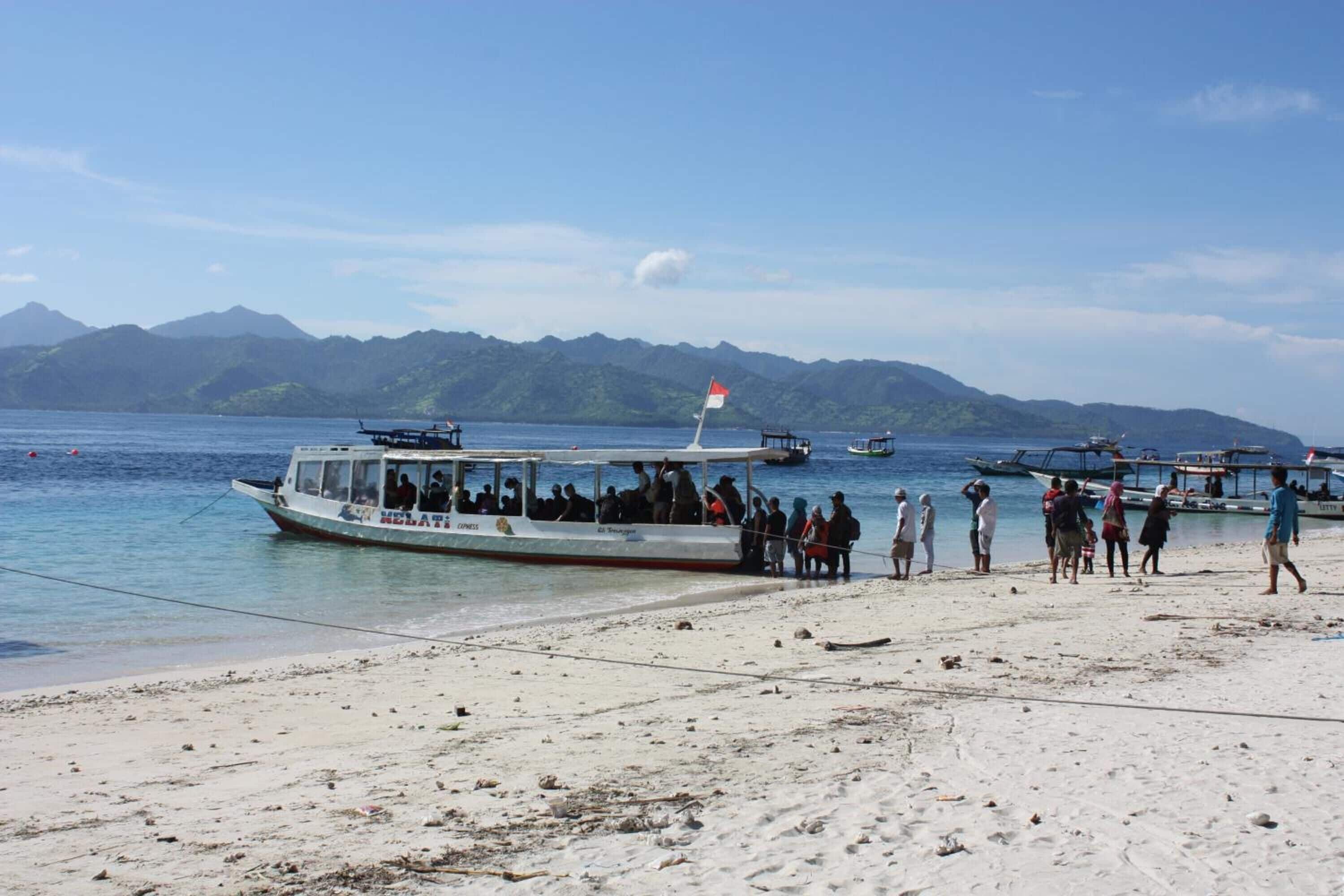 A beach with a boat docked near the shore, people walking on the sand, and mountains in the background.