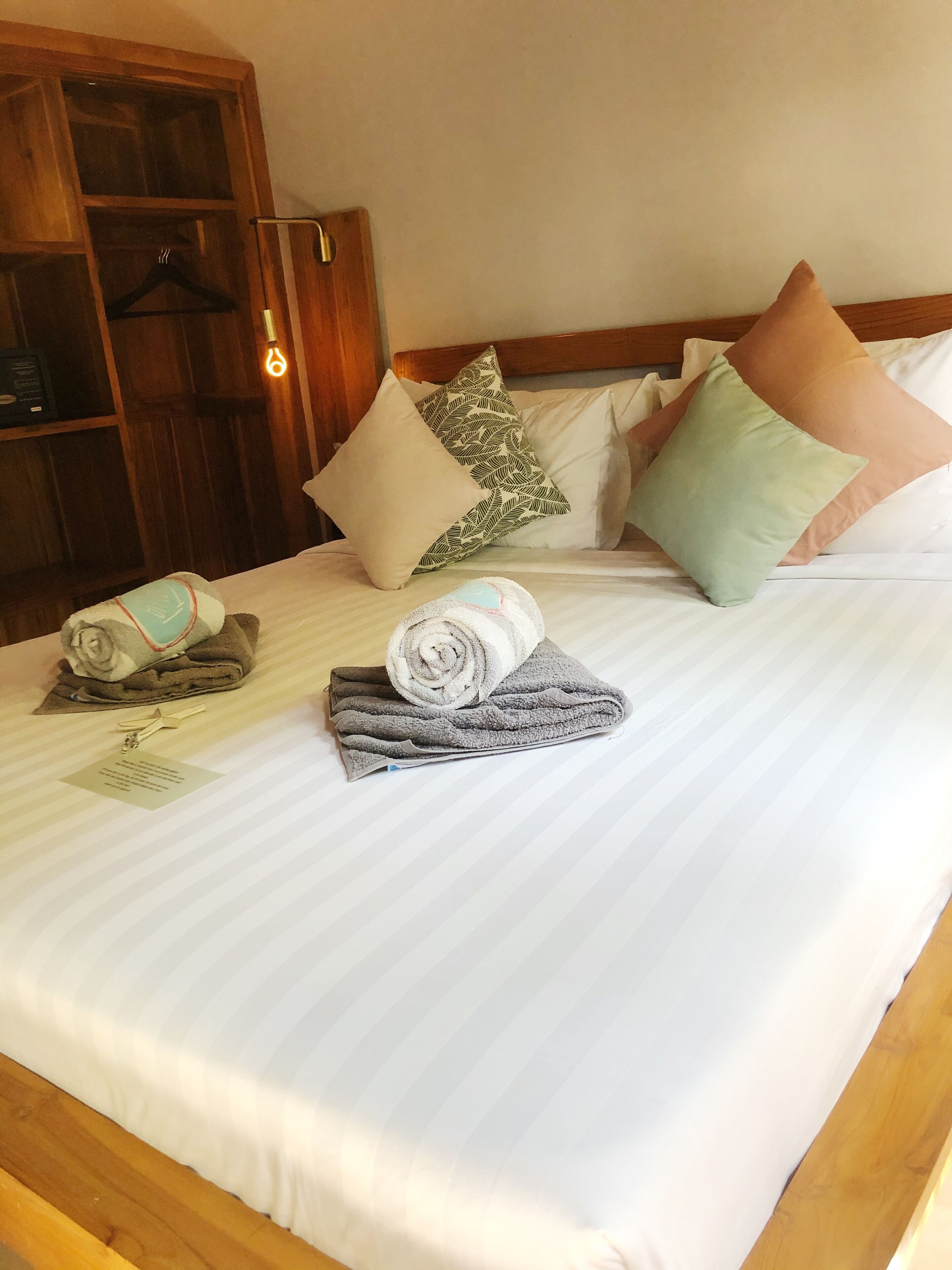 A neatly made bed with rolled towels and a keychain, a wooden wardrobe, and a hanging light fixture.