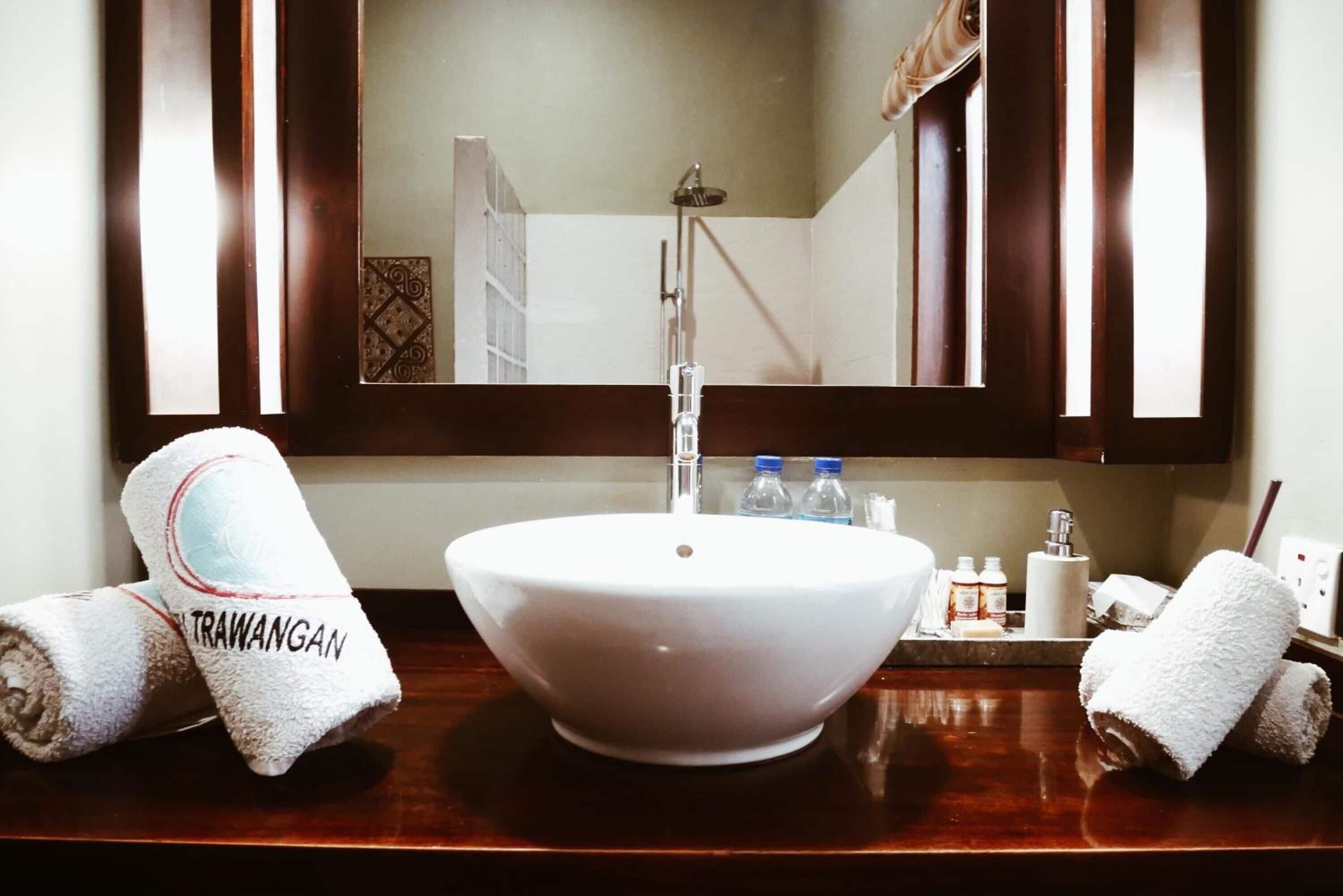A bathroom with a white vessel sink, two towels with 'TRAWANGAN' written on them, a mirror, and toiletries on the counter.
