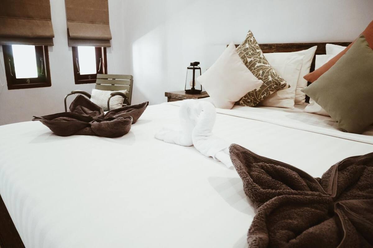 A neatly made bed with white linens, a brown throw blanket, and a brown towel. There are pillows with a leaf pattern and a green pillow. A wooden nightstand with a lantern is visible.