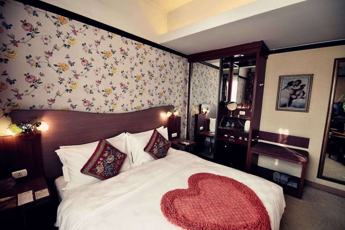 Une chambre d’hôtel avec un grand lit, un papier peint à motifs floraux, un plaid en forme de cœur, un miroir et une table de chevet avec une lampe.