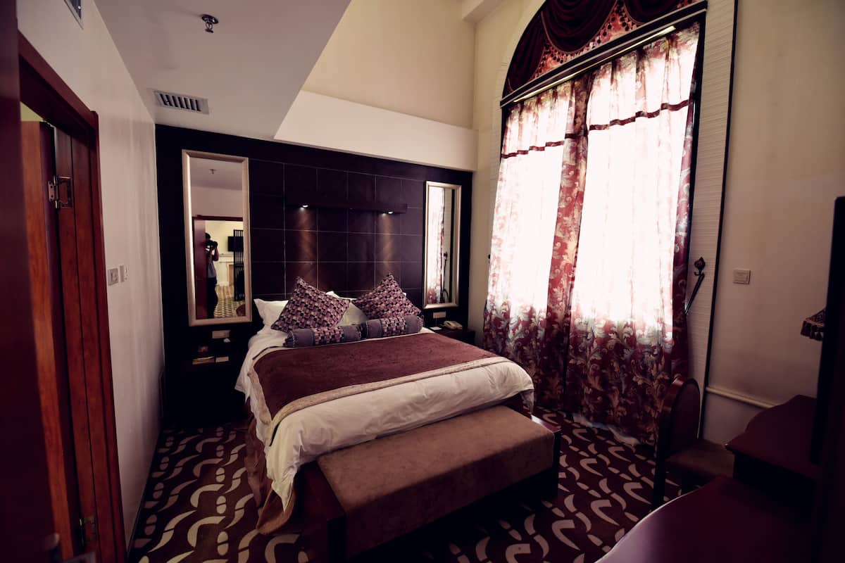 Une chambre avec un grand lit, un banc, un miroir et une fenêtre avec des rideaux.