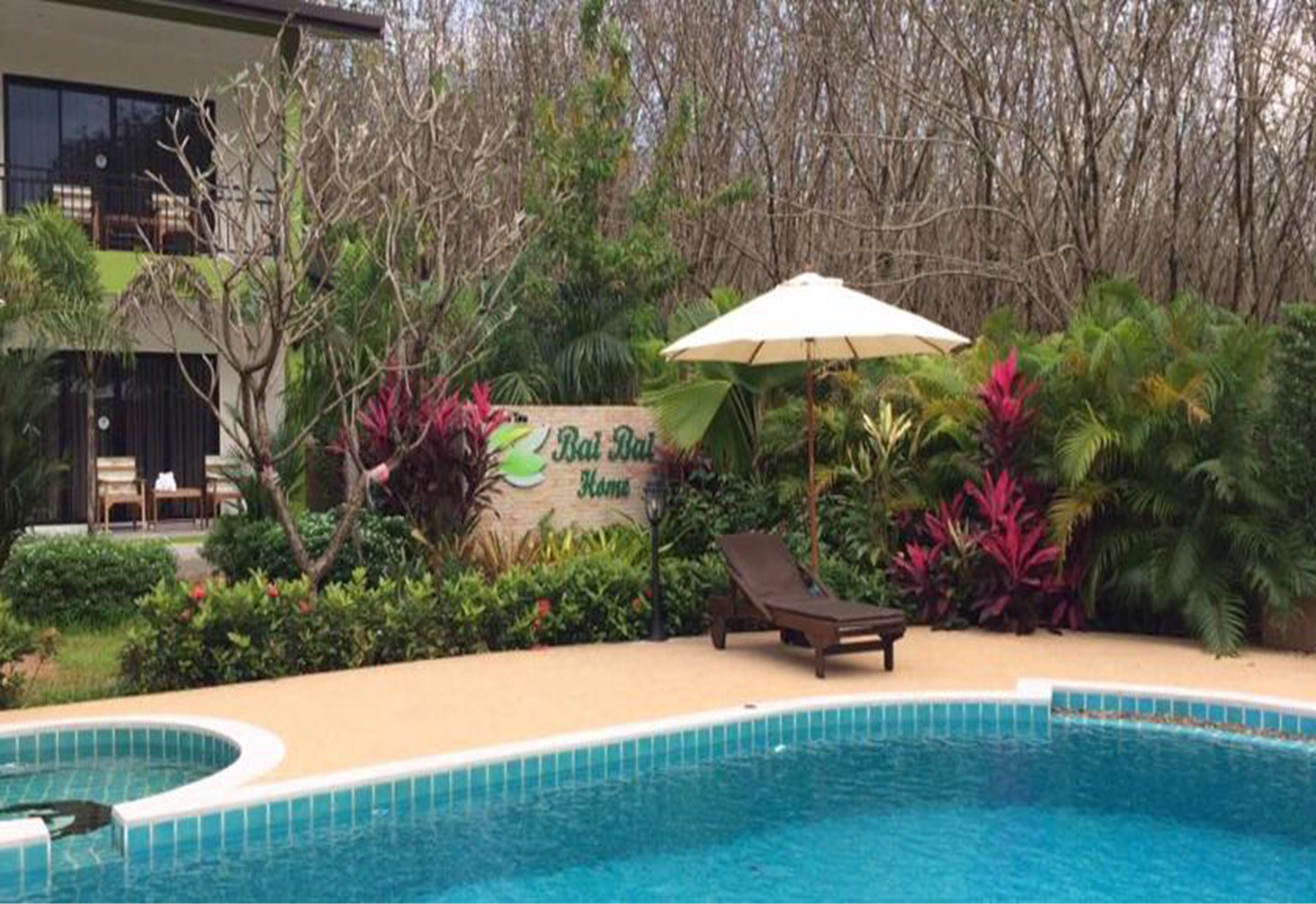Una piscina con una poltrona da spiaggia e un ombrellone, immersa nel verde e con un cartello che recita "Bai Bai Home".