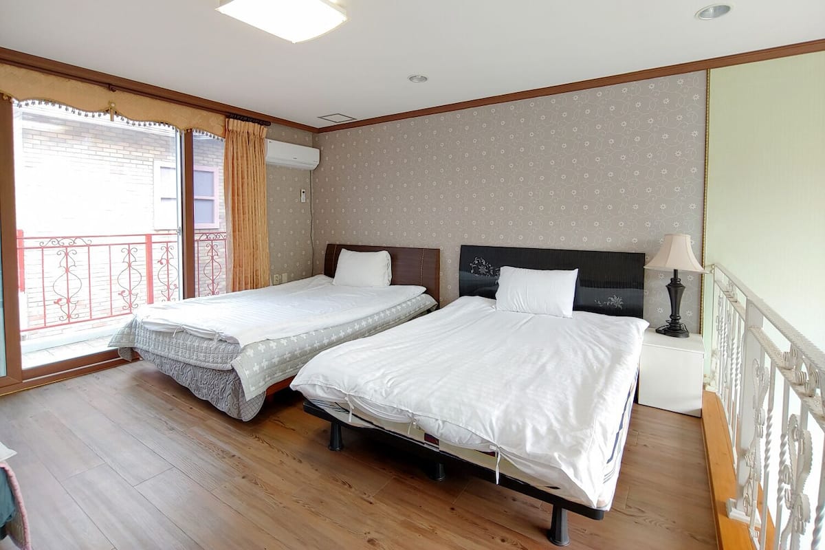 A hotel room with two beds, a balcony with a red railing, and a ceiling light.