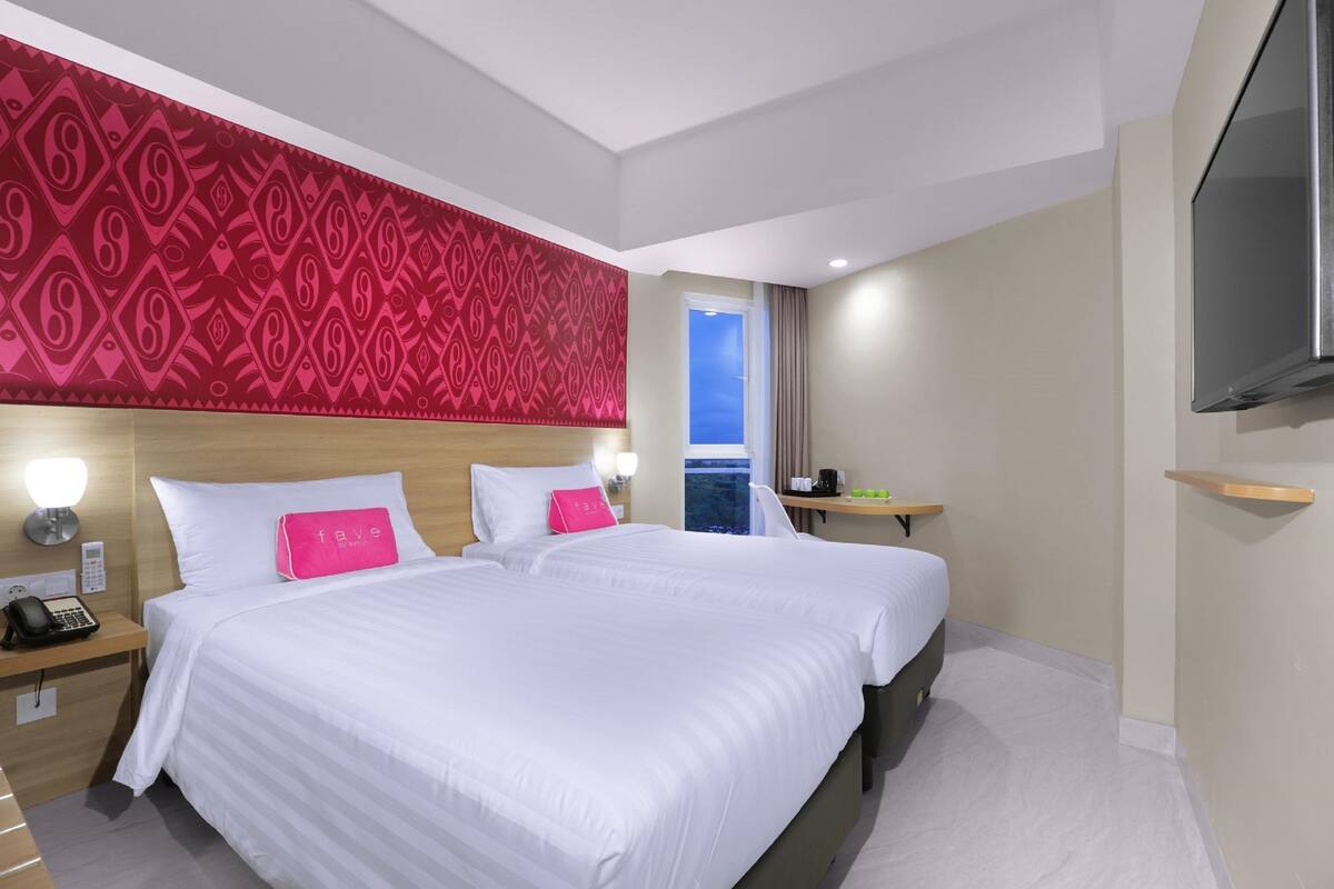 Habitación de hotel con dos camas, un cabecero rojo con estampado, una mesita de noche con teléfono y un televisor montado en la pared.