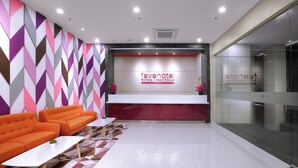 Un área de recepción moderna de hotel con un muro de diseño geométrico, un mostrador de recepción y una zona de estar con sofás naranjas y una mesa blanca.