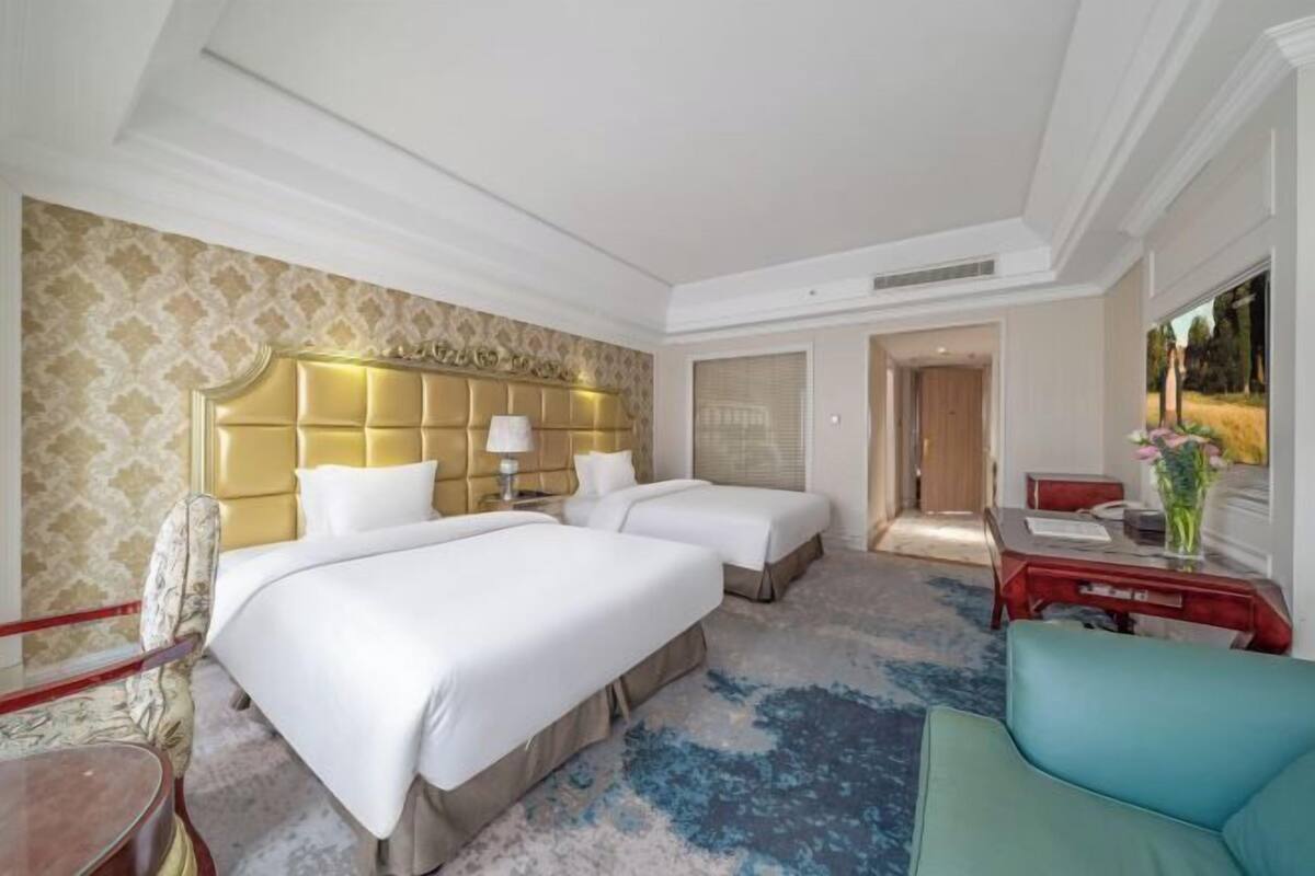 A hotel room with two beds, a large headboard, a desk, a chair, and a view of the outdoors.