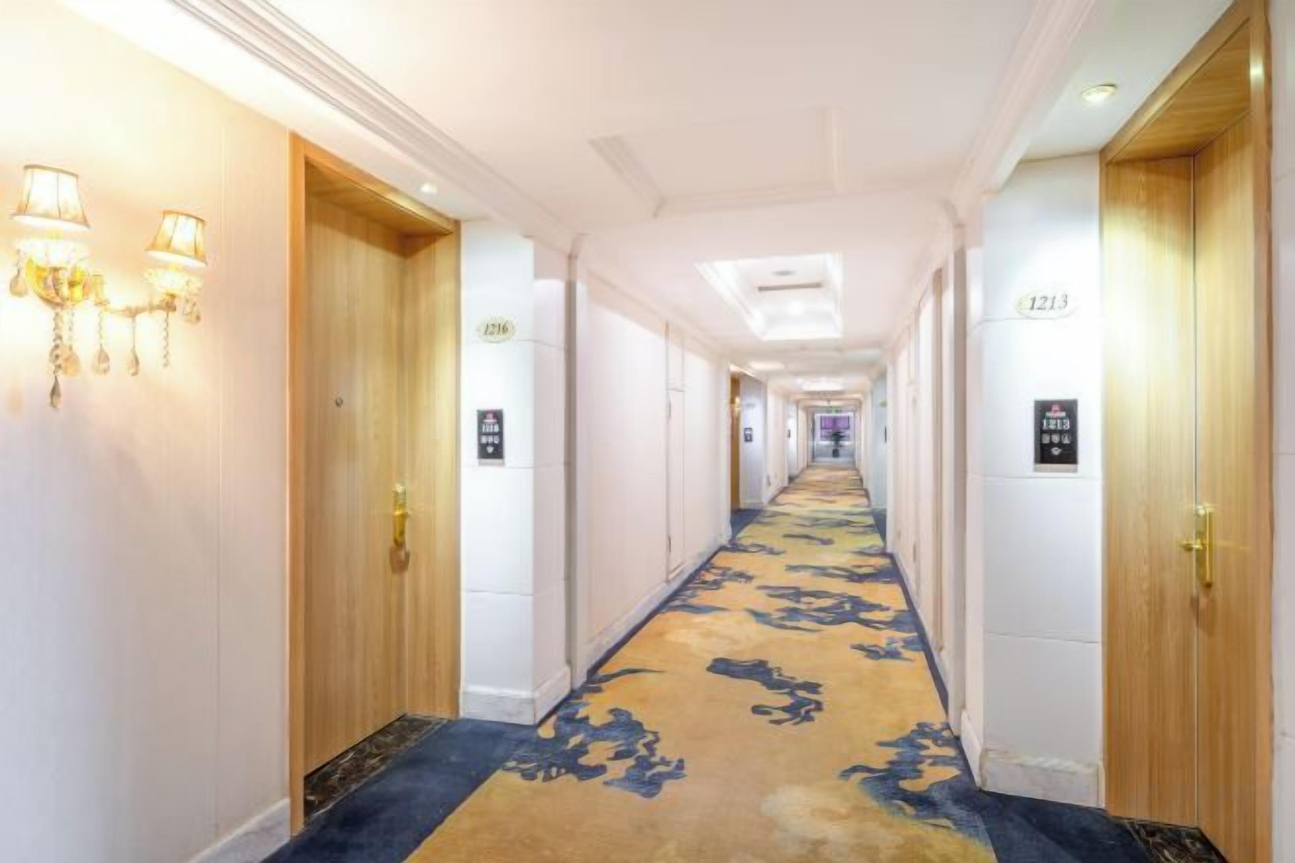 A hotel corridor with a carpeted floor, doors on both sides, and wall-mounted light fixtures.