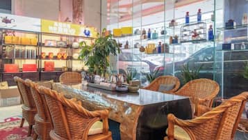 Een hotelhal met een glazen scheiding, rieten stoelen, een marmeren tafel en een plant.