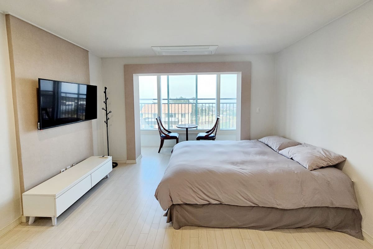 Una habitación de hotel moderna con una cama grande, un televisor de pantalla plana montado en la pared, una pequeña zona de estar con una mesa y sillas, y un balcón con vistas.