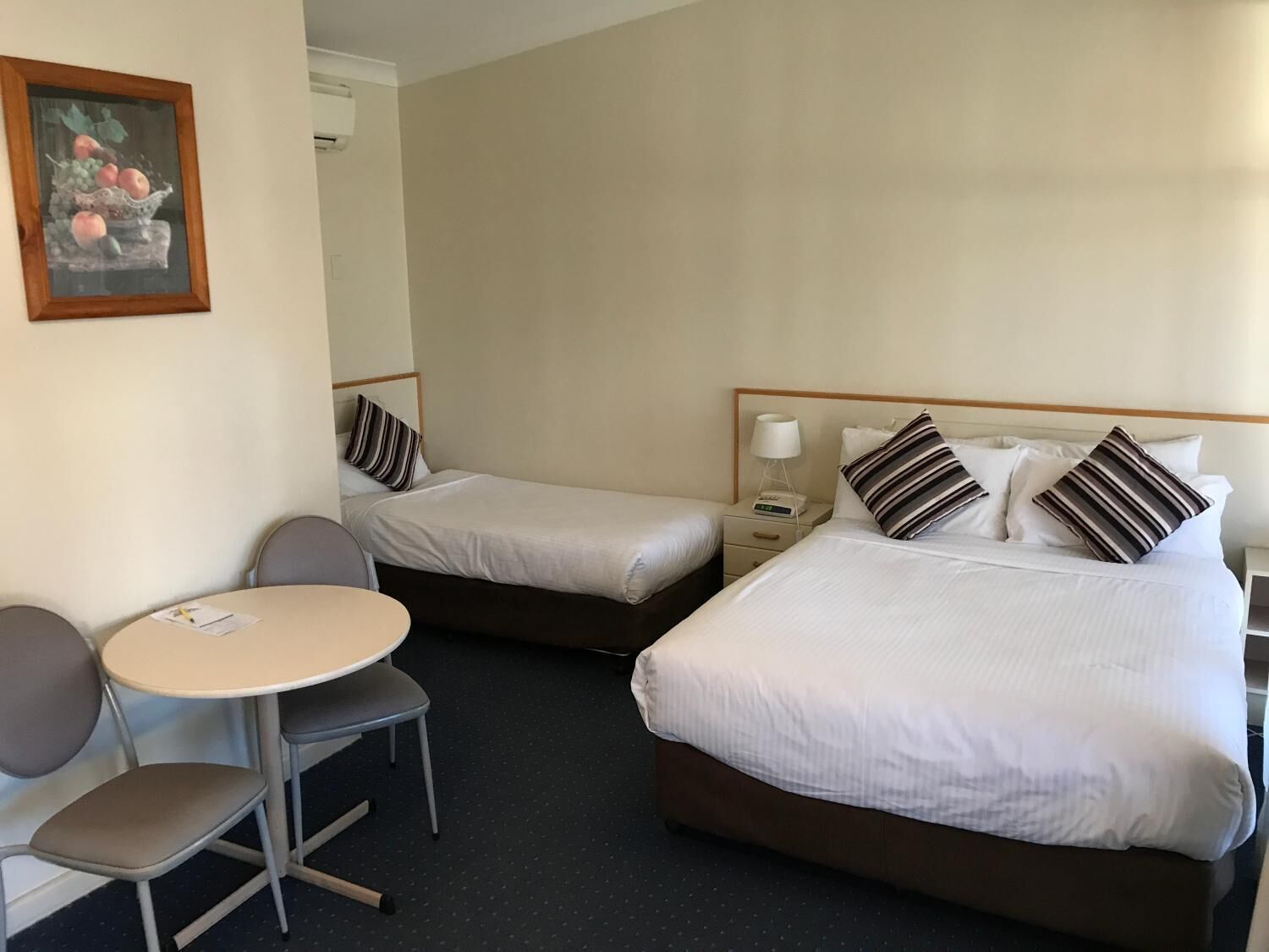 A hotel room with two single beds, a small round table with a chair, and a painting on the wall.