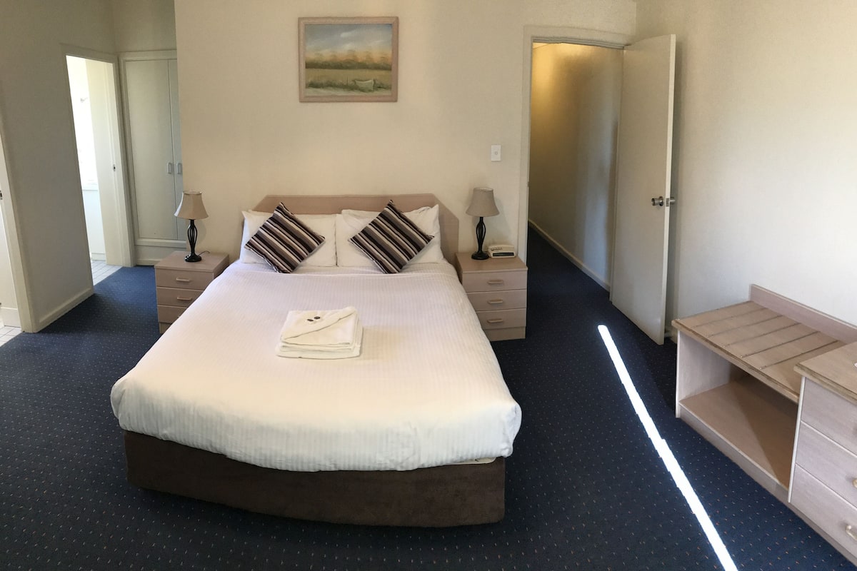 A neatly made bed with a folded towel, two bedside tables with lamps, a painting on the wall, and a door leading to another room.