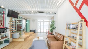 A modern interior with a wooden floor, a brown leather couch, a bookshelf, and a television on a wall. - ALEXA Nimman - Hostel (Chiang Mai)