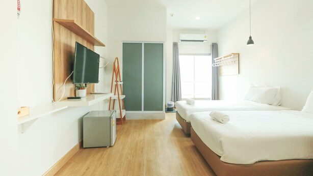 A modern hotel room with a wooden floor, a TV mounted on the wall, a desk with a lamp, a wardrobe, and a bed with white linens. - ALEXA Nimman - Hostel (Chiang Mai)